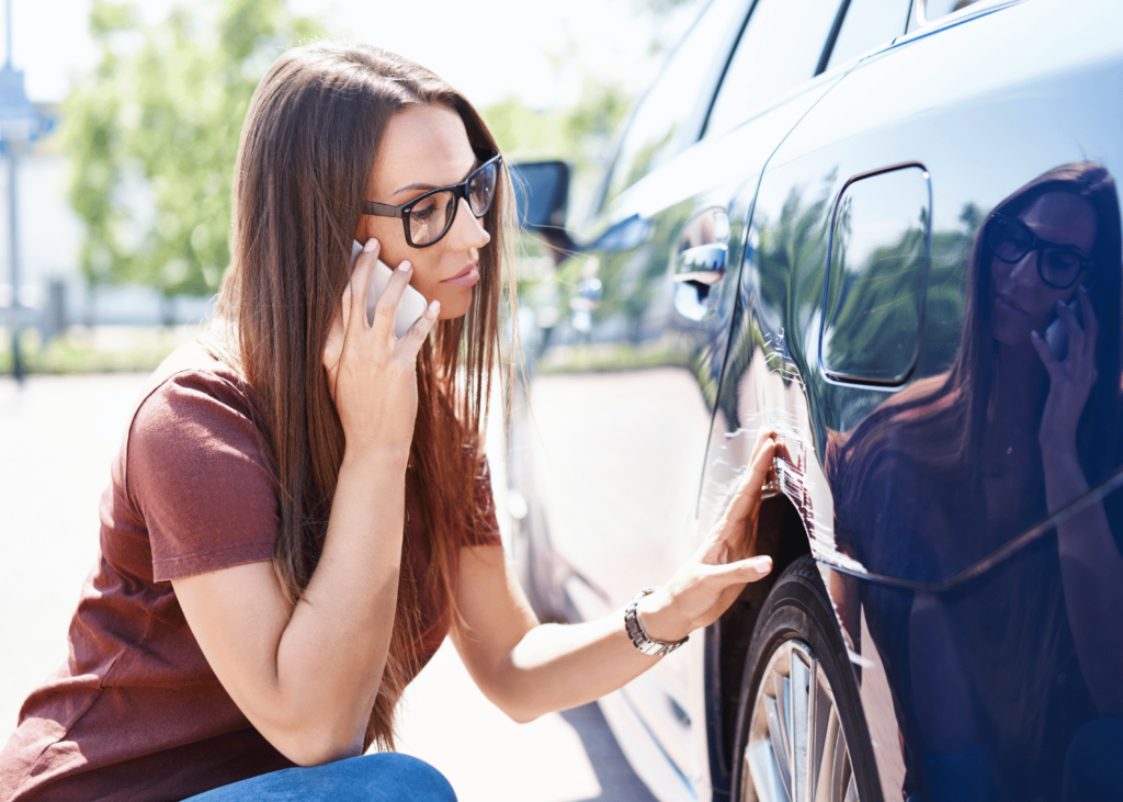 Woman squatting down on mobile phone touching blue car scratch wheel guard in carpark